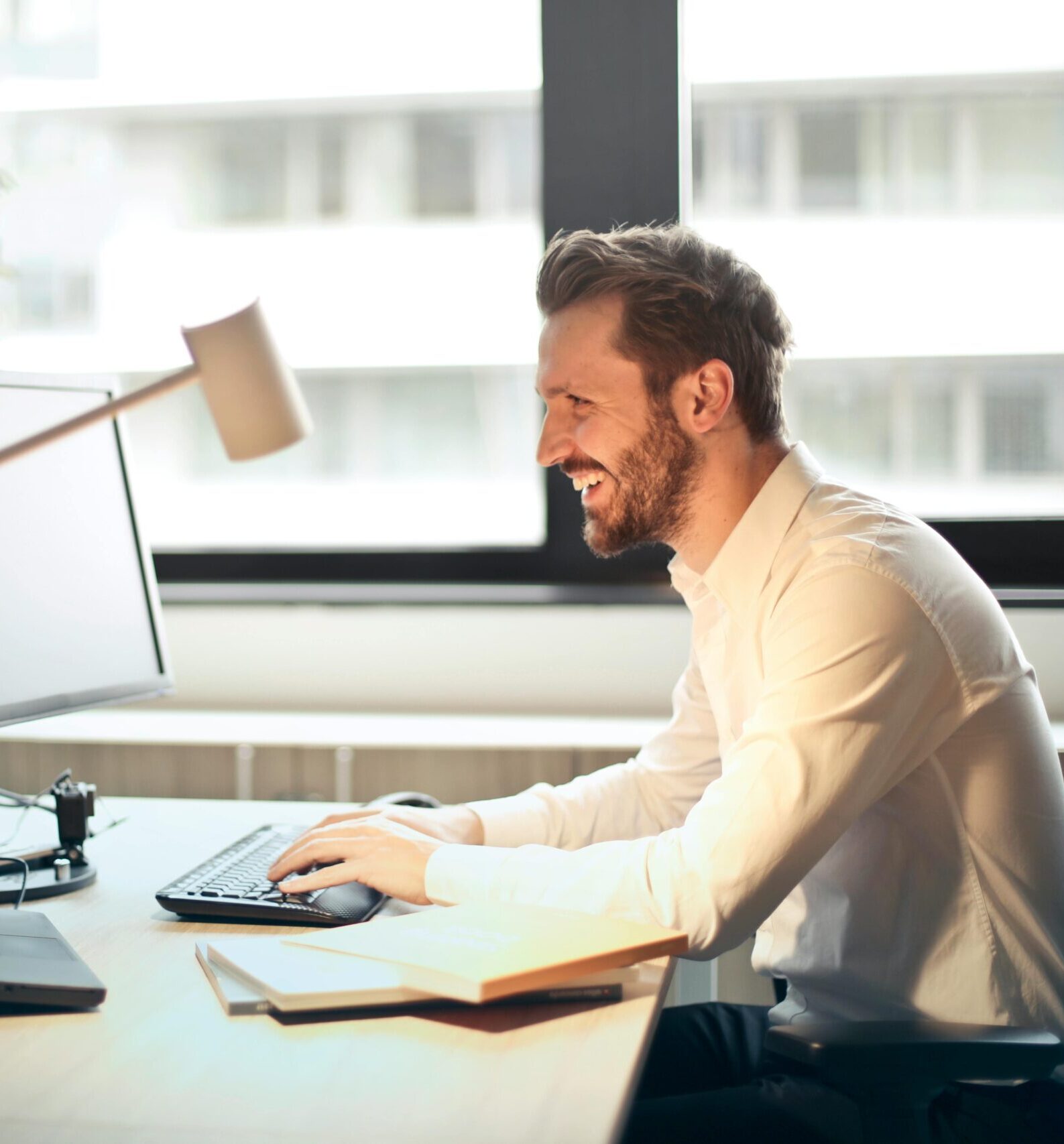 A man smiling while working at an office desk with a computer and natural daylight streaming in through large windows.